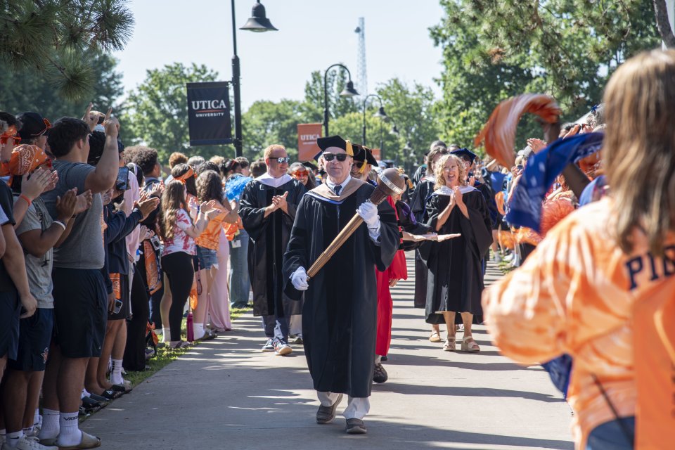 Professor Thomas Rossi acts as Macebearer, leading a procession of faculty down the sidewalk surrounded by cheering students.